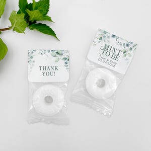 Puede incluir: Dos pequeñas bolsas de regalo blancas con diseños florales verdes y blancos. Una bolsa tiene una etiqueta que dice "Mint to Be" con los nombres Tyler & Emily y la fecha 05.24.2028. La otra bolsa tiene una etiqueta que dice "¡Gracias!"
