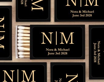 Peut inclure: Boîtes d'allumettes noires avec des lettres dorées et les initiales "NM". Le texte "Nora & Michael June 3rd 2028" est imprimé sur les boîtes. Une boîte est ouverte, révélant les têtes d'allumettes.