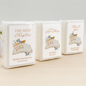 Peut inclure: Trois paquets de serviettes en papier blanc sous emballage plastique transparent. Chaque paquet porte une étiquette illustrée d'un livre ouvert, de lunettes de lecture et de fleurs. Les textes incluent The Next Chapter, Our Love Story et Thank You.