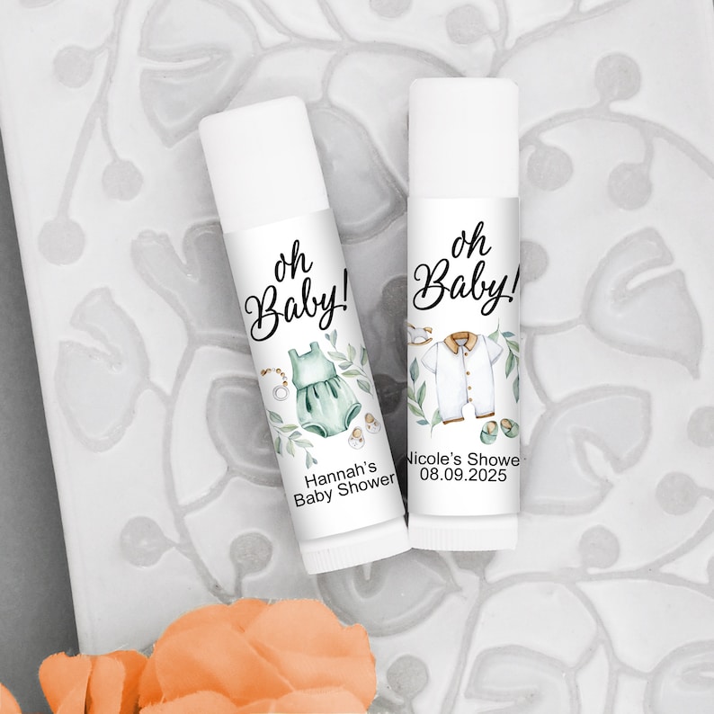 May include: Two white lip balm tubes with baby shower themed labels. The labels feature the text "oh Baby!" and baby-related illustrations, including a dress and a shirt. One label reads "Hannah's Baby Shower".