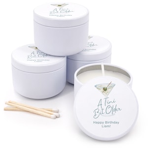 May include: Four white candle tins with a martini glass design and the text "A Tini Bit Older" and "Happy Birthday Liam!"  Three of the tins are closed and one is open with a white candle inside.  There are three wooden matches on the white surface.