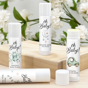 May include: White lip balm tubes with baby shower-themed labels. The labels feature the text "oh Baby!" along with illustrations of a onesie, a car, a moon and stars, and personalized names and dates. The lip balms are displayed on a wooden surface.