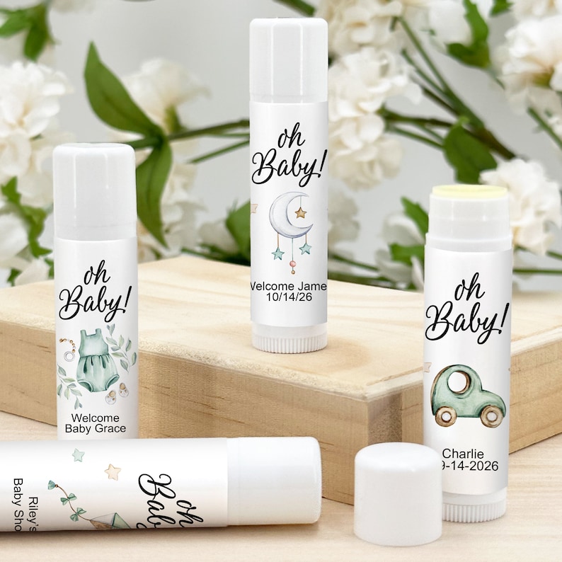 May include: White lip balm tubes with baby shower-themed labels. The labels feature the text "oh Baby!" along with illustrations of a onesie, a car, a moon and stars, and personalized names and dates. The lip balms are displayed on a wooden surface.