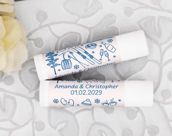 May include: Two white lip balm tubes with light pink labels. The labels feature blue winter-themed illustrations, including skis, a ski lift, and snowflakes. The text "Amanda & Christopher 01.02.2029" is also printed on the labels.