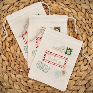 May include: Three off-white drawstring gift bags with red and green postage-themed designs. One bag reads "Deliver To: Whitney" in a red frame. Other stamps include a Santa Claus image and "Do Not Open Until Dec. 25th."