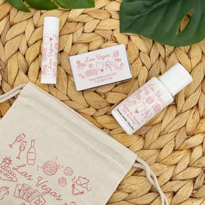 K&ouml;nnte beinhalten: Eine Sammlung von Las Vegas-Themen-Geschenken f&uuml;r Junggesellinnenabschiede. Enth&auml;lt einen Lippenbalsam, Sonnencreme und eine kleine Schachtel, alle mit rosa Text und Illustrationen. Ein Kordelzugbeutel mit Las Vegas-Grafiken ist ebenfalls vorhanden.