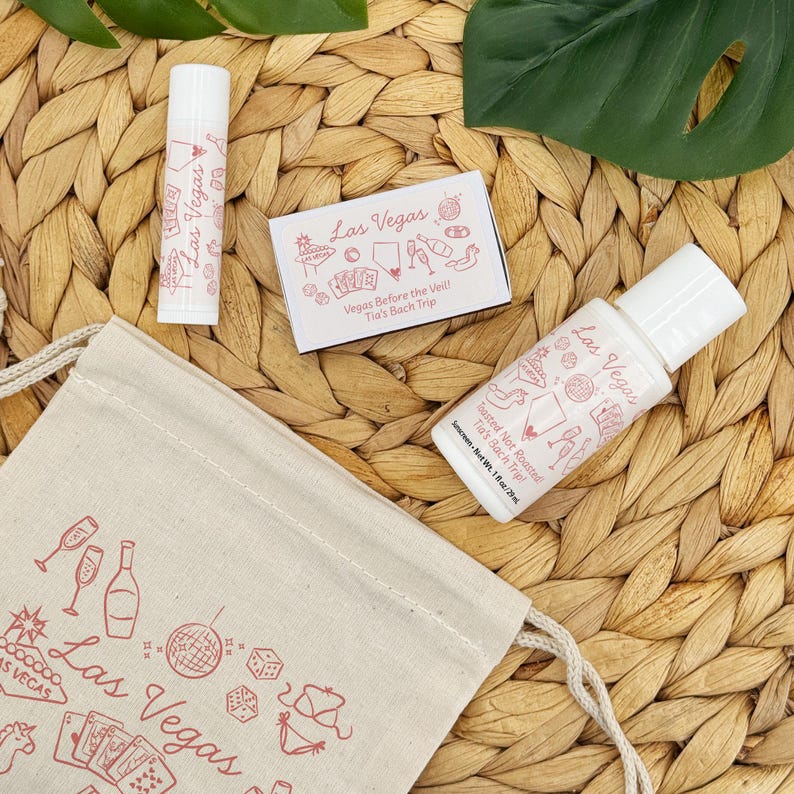 K&ouml;nnte beinhalten: Eine Sammlung von Las Vegas-Themen-Geschenken f&uuml;r Junggesellinnenabschiede. Enth&auml;lt einen Lippenbalsam, Sonnencreme und eine kleine Schachtel, alle mit rosa Text und Illustrationen. Ein Kordelzugbeutel mit Las Vegas-Grafiken ist ebenfalls vorhanden.