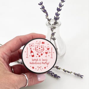 May include: A pink and red phone grip with a heart-shaped cake, wine glasses, candles, and a bottle of wine. The text "Sarah & Ian's Valentine's Party!" is printed on the grip.