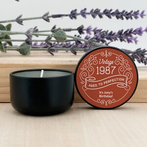 May include: A black candle in a round container with a white wick. The lid, with a brown background, reads "Vintage 1987 Aged to Perfection" and "It's Joey's Birthday!" Decorative lavender sprigs are in the background.