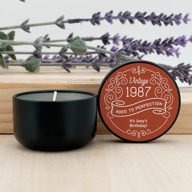 May include: A black candle in a round container with a white wick. The lid, with a brown background, reads "Vintage 1987 Aged to Perfection" and "It's Joey's Birthday!" Decorative lavender sprigs are in the background.