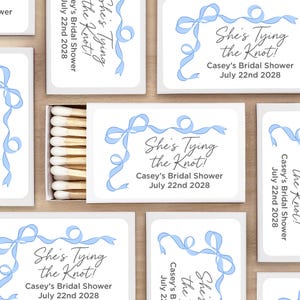 May include: White matchboxes with blue ribbon designs and the text "She's Tying the Knot!" Casey's Bridal Shower July 22nd 2028.