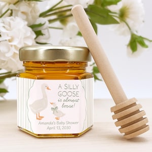 May include: A hexagonal glass jar filled with golden honey, featuring a label with two geese illustrations and the text A SILLY GOOSE is almost loose! Amanda's Baby Shower April 13, 2030. A wooden honey dipper rests beside the jar.