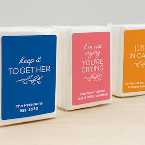 Peut inclure: Trois paquets de mouchoirs avec des étiquettes colorées. L'étiquette bleue dit "keep it TOGETHER" et "The Petersons Est. 2030". L'étiquette rose dit "I'm not crying YOU'RE CRYING" et "Emotions Happen Jess & Will's Wedding". L'étiquette orange dit "JUST IN CASE".