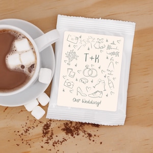 Puede incluir: Un sobre blanco con una tarjeta de color crema en su interior. La tarjeta tiene un diseño dibujado a mano de iconos relacionados con la boda y el texto "T+K Our Wedding!"