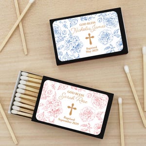 Puede incluir: Dos cajas de cerillas negras con dise&ntilde;os florales y texto personalizado. Una caja presenta flores azules y el texto "God Bless Nicholas James" con una cruz y "Baptized May 2029." La otra caja tiene flores rosas y dice "God Bless Sarah Rose" con una cruz y "Baptized September 2029."