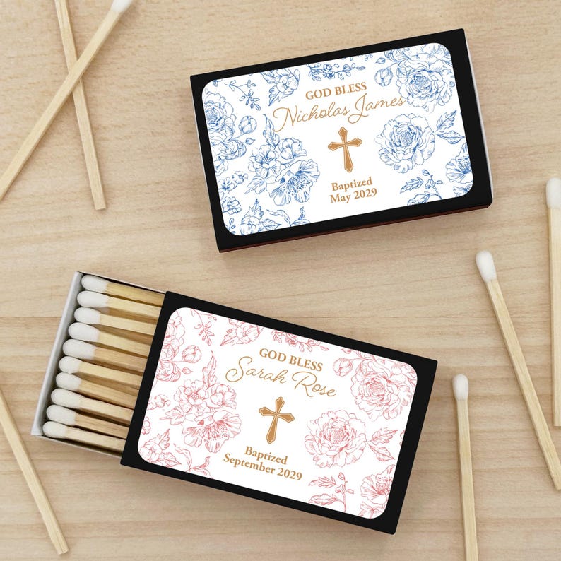 Puede incluir: Dos cajas de cerillas negras con dise&ntilde;os florales y texto personalizado. Una caja presenta flores azules y el texto "God Bless Nicholas James" con una cruz y "Baptized May 2029." La otra caja tiene flores rosas y dice "God Bless Sarah Rose" con una cruz y "Baptized September 2029."