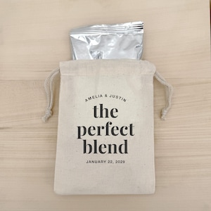以下が含まれることがあります： 「AMELIA & JUSTIN」と「the perfect blend」、日付「JANUARY 22, 2029」の文字が入ったベージュ色の巾着袋。銀色のアルミホイルの小包が入っています。袋は明るい木製の表面に置かれています。