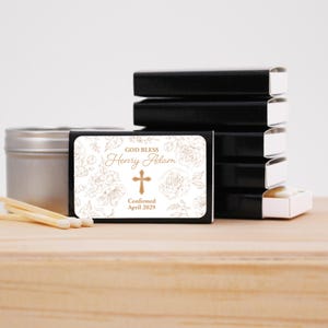 Puede incluir: Cajas de cerillas negras con una etiqueta blanca con dise&ntilde;os florales, una cruz y el texto "GOD BLESS Henry Adam Confirmed April 2029". Tambi&eacute;n se ve una lata plateada y cerillas. Las cerillas est&aacute;n sobre una superficie de madera.