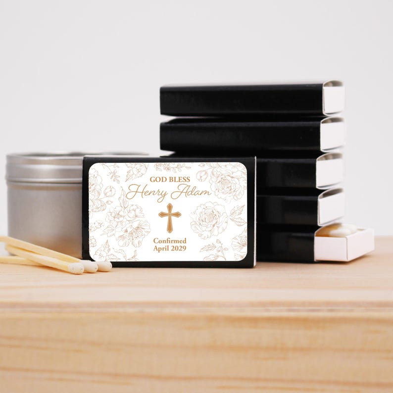 Puede incluir: Cajas de cerillas negras con una etiqueta blanca con dise&ntilde;os florales, una cruz y el texto "GOD BLESS Henry Adam Confirmed April 2029". Tambi&eacute;n se ve una lata plateada y cerillas. Las cerillas est&aacute;n sobre una superficie de madera.