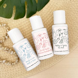 May include: Three white sunscreen bottles with white caps are displayed on a straw hat. Each bottle has a different design with beach-themed illustrations and text. The bottles are labeled "Drunk in Love," "Let's Get Nauti," and "Ashley is Tying The Knot."