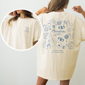 Puede incluir: Camiseta color crema con un gráfico azul en la espalda con botas de vaquero, una guitarra y la palabra "Austin". La parte delantera tiene un pequeño diseño de bota. El texto en la parte posterior dice "Let's Get Rowdy Girls Weekend 2029."