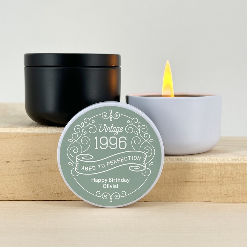 May include: A black candle with a lid, a lit white candle, and a round green label. The label reads "Vintage 1996 Aged to Perfection Happy Birthday Olivia!" in white script. The candles are on a wooden surface.