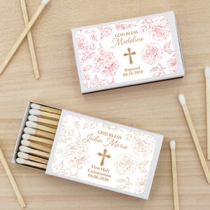 May include: Two white matchboxes with floral designs and personalized text. One reads "God Bless Madeline" with a cross and baptism date. The other says "God Bless Julia Marie" with a cross and First Holy Communion date. Matches are scattered around.