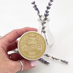 May include: A round, gold and white sticker with the text "Vintage 1989 Aged to Perfection" and "It's Casey's Birthday!" in a decorative font. The sticker is held near a small glass vase with lavender sprigs.