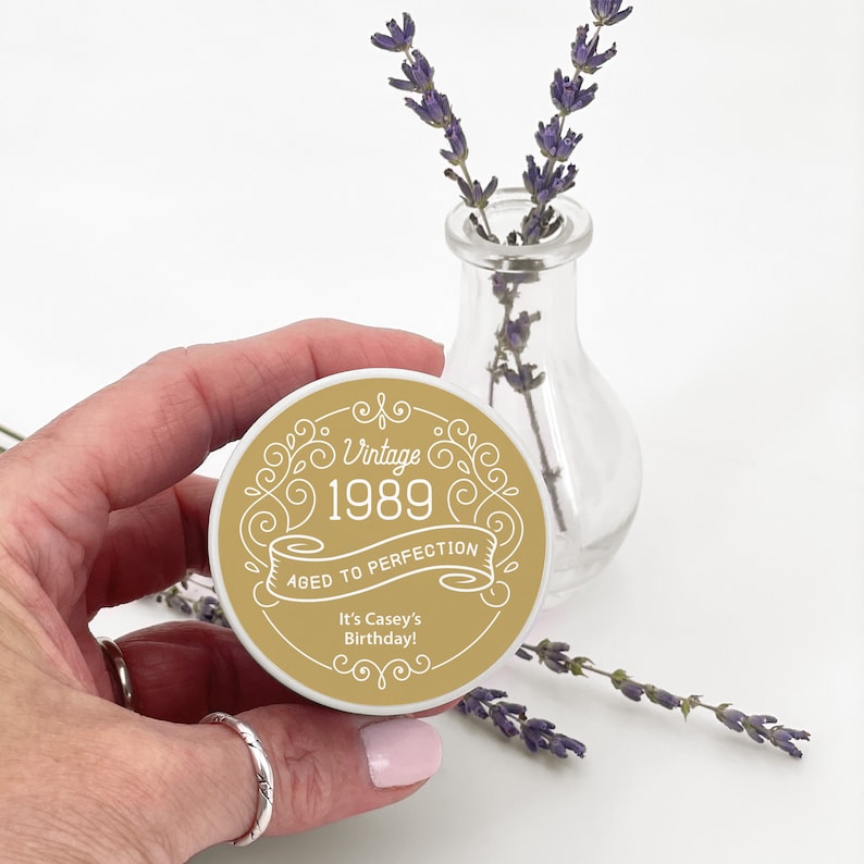 May include: A round, gold and white sticker with the text "Vintage 1989 Aged to Perfection" and "It's Casey's Birthday!" in a decorative font. The sticker is held near a small glass vase with lavender sprigs.