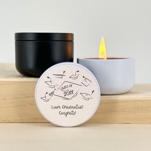 May include: A black candle with a lid, a lit white candle, and a candle lid with graduation-themed graphics and text that reads "Class of 20XX", "Liam Graduated! Congrats!". The candles are on a wooden surface.