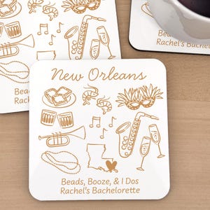 May include: Square white coasters with gold illustrations of New Orleans symbols, such as a saxophone, champagne glasses, and a mask. The text says "New Orleans" and "Beads, Booze, & I Dos Rachel's Bachelorette."