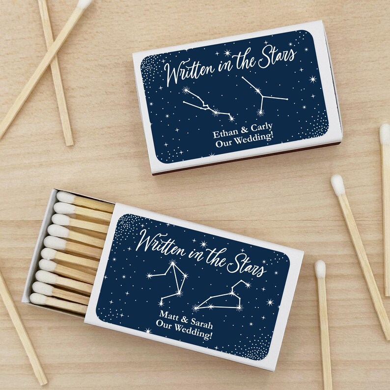 May include: Two white matchboxes with blue and white constellation designs. The text "Written in the Stars" is on each box. One box says "Ethan & Carly Our Wedding!" and the other says "Matt & Sarah Our Wedding!"