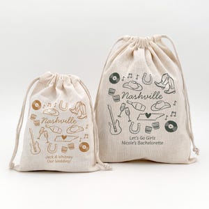 May include: Two beige drawstring bags with Nashville-themed illustrations. The bags feature drawings of cowboy hats, boots, guitars, and records. One bag reads "Jack & Whitney Our Wedding!" and the other says "Let's Go Girls Nicole's Bachelorette".