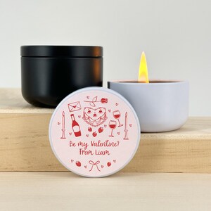 May include: A white candle with a pink lid. The lid has a red heart-shaped cake, wine glasses, a bottle of wine, a rose, and an envelope with the text "Be my Valentine? From Liam".
