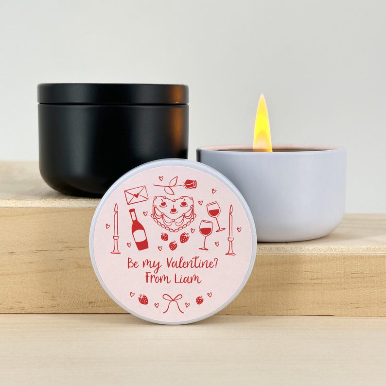 May include: A white candle with a pink lid. The lid has a red heart-shaped cake, wine glasses, a bottle of wine, a rose, and an envelope with the text "Be my Valentine? From Liam".