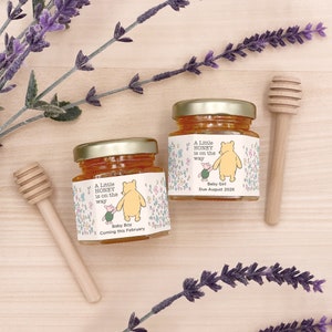 May include: Two small glass jars of honey with Winnie the Pooh and Piglet labels. The labels say "A Little Honey is on the Way" with "Baby Boy Coming this February" and "Baby Girl Due August 2026".