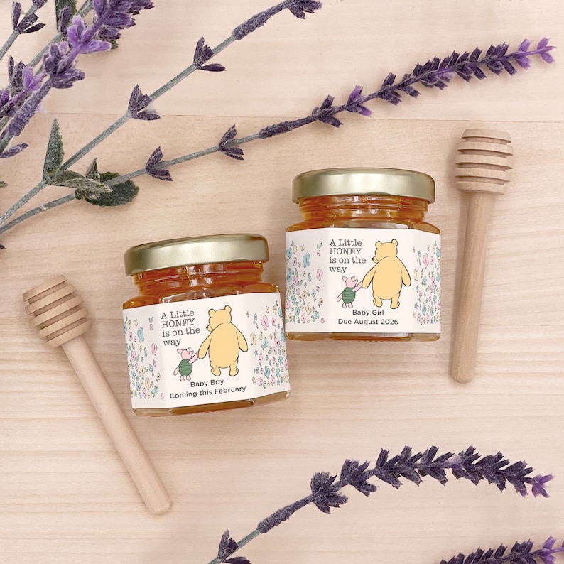 May include: Two small glass jars of honey with Winnie the Pooh and Piglet labels. The labels say "A Little Honey is on the Way" with "Baby Boy Coming this February" and "Baby Girl Due August 2026".