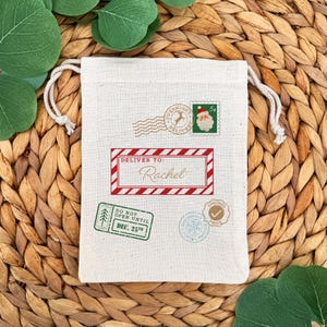 May include: A cream-colored drawstring gift bag with various postage-themed stamps. The bag features a red and white bordered label that reads "Deliver To: Rachel". Other stamps include "Overnight Delivery" and "Do Not Open Until Dec. 25th".