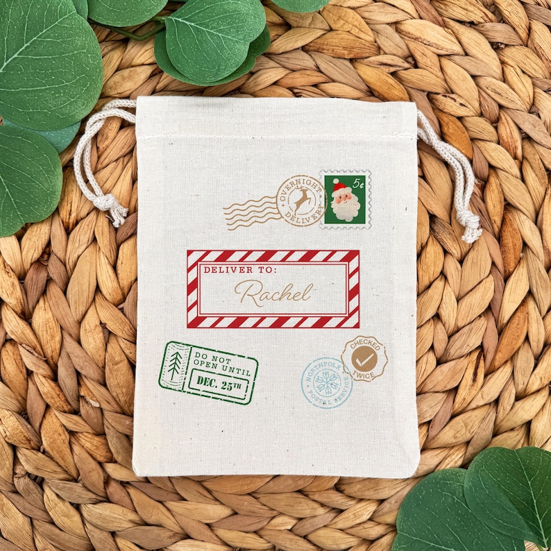 May include: A cream-colored drawstring gift bag with various postage-themed stamps. The bag features a red and white bordered label that reads "Deliver To: Rachel". Other stamps include "Overnight Delivery" and "Do Not Open Until Dec. 25th".