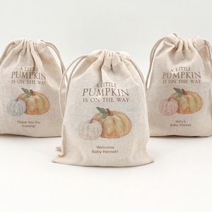 May include: Three beige drawstring bags with pumpkin illustrations and the text "A Little Pumpkin Is On The Way." One bag says "Thank You For Coming!" Another says "Welcome Baby Hannah!" and the third says "Kelly's Baby Shower."