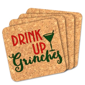 Puede incluir: Un conjunto de cuatro posavasos cuadrados de corcho. Cada posavasos tiene las palabras "DRINK UP Grinches" en letras rojas y verdes, con un gráfico de copa de cóctel verde. Los posavasos tienen un aspecto natural y texturizado.