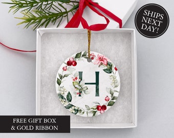 May include: A white ceramic Christmas ornament with a green wreath design and the letter "H" in the center. The wreath is decorated with red berries and green leaves. The ornament is in a white gift box with a red ribbon.