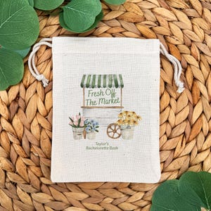 Może przedstawiać: Naturalna pł&oacute;cienna torba ze sznurkiem i akwarelowym wzorem. Projekt przedstawia w&oacute;zek targowy z napisem "Fresh Off The Market" i kompozycjami kwiatowymi. Poniżej znajduje się napis "Taylor's Bachelorette Bash".
