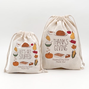 May include: Two beige drawstring bags with Thanksgiving-themed illustrations. One bag says "Let's Get Stuffed" and the other says "Thanks Giving". Both bags feature images of a turkey, pumpkin, and other festive foods. The text "Friendsgiving 2029 Thank You!" is also present.