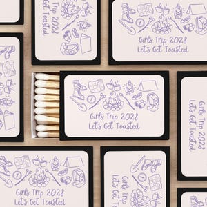 May include: A set of matchboxes with a purple and white design. The matchboxes feature a camping theme with illustrations of a tent, campfire, hiking boots, and a compass. The text on the matchboxes reads "Girls Trip 2028 Let's Get Toasted".