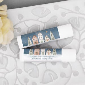 May include: Two white lip balm tubes featuring a blue label with a winter village scene, snow-covered trees, and gold stars. The text reads Amanda & Christopher and Amanda & Christopher Christmas Party 2030 in a dark gray font.