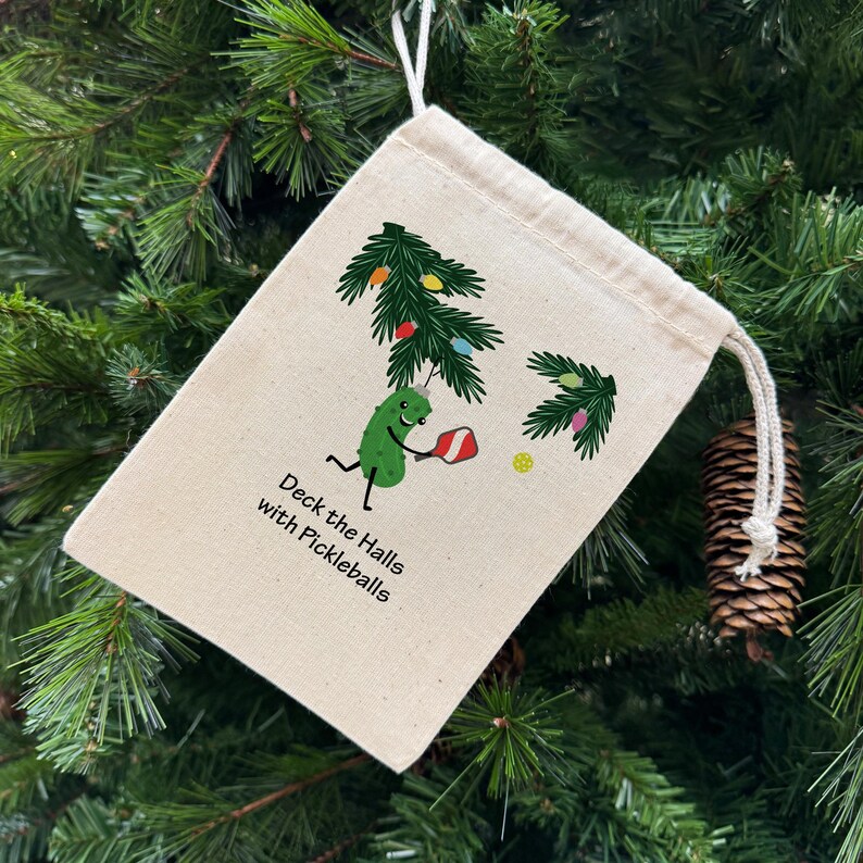 May include: A beige drawstring bag with a festive pickleball-themed design. The bag features a cartoon pickle playing pickleball under a decorated Christmas tree. Text on the bag reads "Deck the Halls with Pickleballs."
