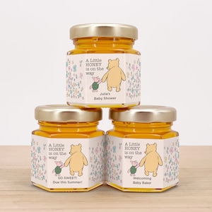 May include: Three small glass jars filled with honey, each with a Winnie the Pooh themed label. The labels feature Winnie the Pooh and Piglet walking together, surrounded by flowers. The text on the labels reads "A Little Honey is on the way". The first label says "Julia's Baby Shower", the second label says "So Sweet! Due this Summer!" and the third label says "Welcoming Baby Baker".