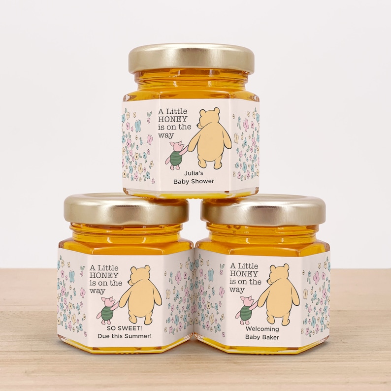 May include: Three small glass jars filled with honey, each with a Winnie the Pooh themed label. The labels feature Winnie the Pooh and Piglet walking together, surrounded by flowers. The text on the labels reads "A Little Honey is on the way". The first label says "Julia's Baby Shower", the second label says "So Sweet! Due this Summer!" and the third label says "Welcoming Baby Baker".