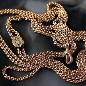 May include: A gold-colored necklace with a delicate chain design. The necklace features a decorative floral clasp with a small turquoise stone and several smaller stones. The chain is draped across a dark, patterned surface.
